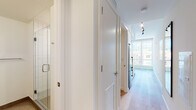 This thoughtfully designed single-floor unit offers a clean, contemporary aesthetic throughout. Light wood flooring runs seamlessly across the open-plan living space, creating a warm and cohesive feel. The compact yet highly functional kitchen is outfitted with stainless steel appliances, including an induction cooktop, built-in oven, microwave, and refrigerator, complemented by crisp white cabinetry, a stainless steel sink with a chrome pull-down faucet, and a distinctive white hexagonal tile backsplash that adds subtle texture to the minimalist design.

The living and sleeping area is cleverly integrated through a partial dividing wall that incorporates a pass-through opening, maintaining an open flow while defining each zone. A built-in platform bed with warm wood-toned under-bed storage drawers adds both style and practicality. Track lighting runs along the ceiling, providing flexible illumination throughout.

A well-appointed hallway connects to the bathroom, which features a glass-enclosed shower with tiled walls. White interior doors and efficient ventilation grilles complete the refined, modern finish of the corridor.

Large floor-to-ceiling windows are a standout feature, flooding the space with natural light and framing expansive urban cityscape views. The overall ambiance is bright, efficient, and contemporary, ideal for modern city living.