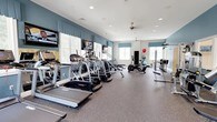 This fitness center offers a thoughtfully designed workout environment with a clean, welcoming atmosphere. The space features blue accent walls contrasted against crisp white ceilings with recessed lighting, creating a bright and energizing ambiance. Large windows with coordinating blue valances line the walls, allowing abundant natural light to fill the room while providing views of the outdoors.

The cardio zone is well-stocked with a row of treadmills, elliptical machines, and upright stationary bikes, including Landice equipment. Multiple flat-screen TVs are wall-mounted throughout, allowing members to stay entertained during workouts. A ceiling fan provides additional air circulation across the space.

Beyond the cardio section, the fitness center includes a strength and functional training area equipped with resistance machines, stability balls, and additional accessories for varied workout routines. The durable speckled flooring throughout is designed to withstand heavy equipment and high-traffic use while providing comfort underfoot.

Overall, the fitness center balances functionality with an aesthetically pleasing design, making it a practical and pleasant space for a full range of exercise activities.