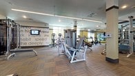 This spacious fitness center is designed to support a comprehensive workout experience across a single, open floor plan. The facility is outfitted with a wide array of cardio equipment, including Matrix treadmills, elliptical trainers, and cycling bikes, all positioned along large windows that invite abundant natural light and offer views of outdoor greenery. The strength training area features multiple cable machines, weight benches, and selectorized resistance equipment arranged to maximize accessibility and floor space. A dedicated group fitness and studio room is enclosed by full-length glass walls and mirrors, housing spinning bikes, medicine balls, stability balls, resistance bands, and exercise mats — ideal for classes or individual functional training. Full-length mirrors line multiple walls throughout the facility, visually expanding the space and aiding in form correction. The interior design features yellow accent columns, a teal-toned studio backdrop, and patterned multi-tone carpet tiles throughout. A standout design element is the motivational typography wall, displaying words such as "inspire," "speed," "deeply," and "results," reinforcing an energizing atmosphere. Recessed and linear LED lighting ensures consistent, bright illumination across the entire floor.