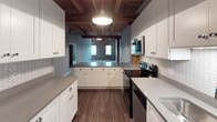This single-floor loft-style unit masterfully combines industrial heritage with contemporary design. The defining architectural feature throughout is the exposed wood beam ceiling system, with rich, warm-toned timber beams and planking that lend character and a sense of height to every room. The open living area is generously proportioned, with plush carpeting, soft grey walls, and a striking original brick accent wall that reinforces the space's historic industrial roots. A structural wood post adds to the authentic loft aesthetic.

The kitchen is well-appointed and highly functional, featuring white shaker-style cabinetry with ample storage, grey quartz countertops, a herringbone tile backsplash, hardwood-style flooring, and a full suite of stainless steel appliances including a range, microwave, dishwasher, and refrigerator. A walk-in pantry provides additional storage convenience.

The bedrooms are carpeted and quiet, each benefiting from the distinctive beam ceiling overhead. One bedroom features a skylight that draws natural light into the space, while another includes a window with exterior views. Both bedrooms offer walk-in closet space with built-in shelving and hanging rods.

The bathroom is clean and modern, finished in white subway tile surrounding a tub-shower combination, complemented by grey quartz dual vanity countertops, white cabinetry, and large-format floor tile.