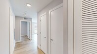 This single-floor space presents a clean, modern aesthetic defined by light hardwood flooring, neutral gray walls, and crisp white trim that flows consistently throughout. The open-concept kitchen is well-appointed with white shaker cabinetry, a subway tile backsplash, stainless steel appliances including a range with microwave, and a full-size refrigerator — all set against a bright, airy backdrop. A large window in the kitchen and dining area allows generous natural light to pour in, offering views of the surrounding neighborhood.

The living areas are generously sized, with multiple rooms featuring floor-to-ceiling windows fitted with horizontal blinds, inviting abundant sunlight and a sense of openness. The hallway connecting the rooms is well-lit with recessed ceiling fixtures and includes multiple interior doors, providing clear separation between spaces.

The bathroom features a marble-style tile floor, clean white walls, a tiled shower, and brushed nickel grab bars, reflecting a practical yet refined design. Throughout the unit, the consistent use of light tones, hardwood flooring, and modern fixtures creates a cohesive, contemporary ambiance that feels fresh and move-in ready.