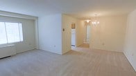 This single-floor space presents a clean, understated interior designed for comfortable everyday living. The main living area is finished with wall-to-wall beige carpeting that extends throughout, creating a warm, cohesive feel underfoot. Smooth white and cream-toned walls keep the space feeling bright and neutral, providing a versatile backdrop for a variety of furnishing styles.

A partial dividing wall separates the living area from the adjoining dining space, offering a degree of definition between the two zones while maintaining an open, connected flow. The dining area is anchored by an elegant chandelier, adding a touch of classic detail to the otherwise simple aesthetic.

Beyond the dining space, the kitchen is visible and features a white refrigerator, with cabinetry overhead providing practical storage. A hallway leads further into the unit, suggesting additional rooms such as bedrooms or a bathroom beyond the main living areas.

Overall, the space maintains a straightforward, move-in-ready character with consistent finishes throughout — neutral carpeting, painted walls, and traditional light fixtures — making it a functional and adaptable living environment.