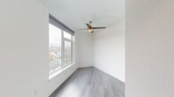 Located at 2180 NE 47th Ave in Portland, Oregon, this efficiently designed studio unit offers a thoughtful single-floor layout that maximizes every inch of its compact footprint. The combined living, dining, and kitchen area is anchored by light gray laminate wood flooring that flows seamlessly throughout the space, creating a sense of visual continuity and warmth.

The kitchen is a standout feature, outfitted with contemporary gray flat-front cabinetry — both upper and lower — paired with clean white countertops. It includes a built-in electric cooktop, an under-counter microwave, a refrigerator, a single-basin sink, and open display shelving, providing efficient storage and functionality in a streamlined design.

The living area benefits from large windows fitted with roller shades, allowing generous natural light to fill the room while framing views of the surrounding Portland neighborhood rooftops and treeline. A ceiling fan with a light fixture adds both comfort and a practical design element overhead.

The bathroom is clean and bright, finished in white with wood-look flooring, and features a shower/tub combination, toilet, and an accessible grab bar, reflecting a modern, utilitarian design. The overall aesthetic of the unit is crisp and contemporary, with neutral tones and modern finishes throughout.