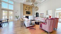 Located at 103 Inspiration Ave in Bluffton, South Carolina, this single-floor clubhouse at Mystic Bluff Apartment Homes is a thoughtfully designed community gathering space that blends coastal charm with modern elegance. Soaring coffered and tray ceilings define the interior throughout, creating an airy, grand atmosphere that is immediately felt upon entering the welcoming foyer. The entry features warm hardwood-style flooring, an ornate area rug, and plentiful natural light pouring in through transom windows and glass-paneled double doors. The open lounge area serves as the heart of the space, anchored by a striking textured stone fireplace wall with a mounted television, complemented by plush neutral sofas, vibrant crimson accent chairs, and sculptural driftwood-inspired pendant chandeliers. Adjacent to the lounge, a beautifully appointed kitchen showcases crisp white cabinetry, grey subway tile backsplash, stainless steel appliances, and a substantial waterfall-edge island illuminated by amber glass pendant lights — ideal for community events. A separate dining area enveloped in floor-to-ceiling windows and elegant draping offers an abundance of natural light and views of the surrounding community, featuring rustic wood dining tables beneath wrought-iron chandeliers. Warm hardwood-style flooring flows seamlessly throughout, tying together each distinct area into a cohesive, inviting whole.