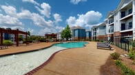 This outdoor pool area in Oxford, Mississippi offers a well-appointed, resort-style aquatic retreat designed for relaxation and recreation. The centerpiece is a generously sized freeform swimming pool with a distinctive curvilinear shape, trimmed with classic brick coping and featuring a gradual zero-entry shallow end that transitions into deeper water. A separate circular hot tub is positioned nearby, providing a dedicated space for relaxation. The expansive surrounding sun deck is finished in a warm aggregate concrete, offering ample room for lounging and movement. Multiple chaise lounge chairs and seating arrangements are distributed across the deck, accommodating a variety of leisure activities. Two striking red-stained wooden pergolas anchor the seating areas, adding architectural character and shade to the space. The perimeter of the pool area is enclosed by a black iron fence, with lush ornamental grasses and mature trees providing a natural privacy buffer. The surrounding multi-story residential buildings, clad in a combination of vinyl siding and brick, are visible from the pool area, reinforcing the community-oriented setting. Under the open Mississippi sky, the space is bathed in natural light, creating a vibrant and inviting atmosphere ideal for outdoor enjoyment throughout the warmer months.