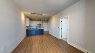 This well-appointed single-floor residence showcases a thoughtfully designed open-concept layout that seamlessly connects the living area and kitchen. The living space features warm wood-look luxury vinyl plank flooring, neutral light-gray walls, and track lighting that provides both ambient and accent illumination. Large windows along the exterior wall allow natural light to fill the room, creating a bright and welcoming atmosphere.

The kitchen is a standout feature, equipped with stainless steel appliances including a refrigerator, electric range, and over-the-range microwave. White upper cabinetry contrasts elegantly with dark wood-grain lower cabinets, complemented by light quartz countertops and a subtle tile backsplash. A bold navy-blue kitchen island with counter seating adds a striking design element and additional workspace, while a matte black faucet and undermount sink complete the modern aesthetic.

The bedrooms are finished with plush neutral carpet, offering comfort and quiet, and feature closet access and natural light from windows with horizontal blinds. The bathroom is clean and functional, featuring a tub-shower combination with sliding glass doors, wood-look flooring, and a contemporary vanity with matte black hardware that ties together the unit's cohesive modern design language.