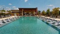 Located at 1045 Williamsburg Way in Evans, Georgia, this single-level outdoor amenity space is designed to serve as a premier recreational hub within a contemporary residential community. At its centerpiece is a generously sized resort-style swimming pool with crystal-clear blue water, flanked by rows of sleek lounge chairs ideal for relaxing under the Georgia sun. Shaded umbrella seating is positioned poolside for added comfort.

Adjacent to the pool is a covered pavilion featuring a warm wood-paneled ceiling, ceiling fans, and an open-air design that invites outdoor living. Inside the pavilion, a well-appointed utility area showcases teal cabinetry with white countertops, a decorative geometric tile backsplash, and vintage-style pendant lighting — combining functionality with design-forward style.

The surrounding grounds include a dedicated outdoor grilling station with brick and wood privacy screening, thoughtfully integrated into lush landscaping featuring ornamental grasses, mulched beds, and young shade trees. Concrete walkways connect the various areas seamlessly, while classic lamp posts and brick accents add architectural character. Views of the broader multi-story residential community frame the space, reinforcing its role as a vibrant communal gathering destination.