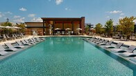 Located at 1045 Williamsburg Way in Evans, Georgia, this single-level outdoor amenity space is designed to serve as a premier recreational hub within a contemporary residential community. At its centerpiece is a generously sized resort-style swimming pool with crystal-clear blue water, flanked by rows of sleek lounge chairs ideal for relaxing under the Georgia sun. Shaded umbrella seating is positioned poolside for added comfort.

Adjacent to the pool is a covered pavilion featuring a warm wood-paneled ceiling, ceiling fans, and an open-air design that invites outdoor living. Inside the pavilion, a well-appointed utility area showcases teal cabinetry with white countertops, a decorative geometric tile backsplash, and vintage-style pendant lighting — combining functionality with design-forward style.

The surrounding grounds include a dedicated outdoor grilling station with brick and wood privacy screening, thoughtfully integrated into lush landscaping featuring ornamental grasses, mulched beds, and young shade trees. Concrete walkways connect the various areas seamlessly, while classic lamp posts and brick accents add architectural character. Views of the broader multi-story residential community frame the space, reinforcing its role as a vibrant communal gathering destination.