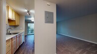 This single-floor residence is organized into a practical, functional layout that makes efficient use of its space. The galley-style kitchen is positioned adjacent to the main living area and features light wood cabinetry, white appliances including a refrigerator and dishwasher, a countertop with a sink, and smooth white countertops. Track lighting overhead and a view directly into the living area give the kitchen an open, connected feel.

The living area is generously proportioned, finished with neutral-toned carpet and painted in soft gray tones. A large sliding glass door fitted with vertical blinds fills the space with natural light and provides direct access to an outdoor balcony, offering views of surrounding greenery. A centrally located electrical panel is mounted on a dividing wall between the living and adjoining rooms.

Additional rooms, visible in the floor plan scan, include carpeted bedroom spaces with gray accent walls and ample room for furnishings. The full bathroom is equipped with a tub-shower combination featuring white tile surround, a wood-vanity with storage below, a toilet, and durable wood-look vinyl flooring. Throughout the unit, clean white baseboards and neutral wall tones contribute to a consistent, understated aesthetic.