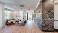 Located at 339 Boston Post Rd in Marlborough, Massachusetts, this single-floor VESA amenity and leasing suite spans a generous footprint designed to balance professional function with residential comfort. The entry lobby immediately sets an elevated tone, anchored by a striking floor-to-ceiling feature wall clad in geometric, chevron-patterned wood tiles backlit to highlight its intricate texture, with the VESA brand name prominently displayed in illuminated lettering. Light oak hardwood flooring runs throughout, creating warmth and visual continuity across the space. Large black-framed glass doors and floor-to-ceiling windows flood the entry and lounge areas with abundant natural light and provide views to the surrounding landscape. A welcoming lounge area is furnished with contemporary seating, layered rugs, and curated wall art, offering a comfortable setting for residents and visitors alike. A separate lounge room features a dramatic teal ceiling, pendant lighting, and oversized windows dressed in sheer curtains, creating an intimate yet airy atmosphere. The space also includes a snack and refreshment station with open shelving, a dedicated mailroom with built-in mailboxes and display shelving, a private glass-walled office, and a spacious storage or package room lined with wall-mounted shelving and durable hardwood floors. A long corridor connects all areas efficiently, completing this well-appointed community hub.