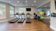 Located at 8820 Autumn Woods Drive in Raleigh, NC, this single-story fitness facility is thoughtfully designed to accommodate a comprehensive range of workout activities across multiple distinct zones. The layout, as revealed by the floor plan overview, is expansive and efficiently organized to maximize usable floor space.

The cardio area features rich hardwood-style flooring and is bathed in natural light from large, floor-to-ceiling windows and a wraparound bay window configuration. Multiple treadmills, elliptical trainers, and stationary bikes are positioned to take advantage of the outdoor views, while ceiling fans maintain airflow throughout the space.

A dedicated free weights zone features durable rubber flooring, a full rack of dumbbells, and large wall-mounted mirrors that visually expand the room. A wall-mounted flat-screen television provides entertainment during workouts.

The strength training area offers additional rubber flooring for safety, cable machines, a weight bench, and selectorized equipment, all set against floor-length mirrors and windows that bring in natural light. Recessed lighting throughout each zone ensures consistent, well-distributed illumination. Medicine balls, resistance equipment, and stability accessories round out the facility's offerings, making this a versatile and well-appointed fitness destination.