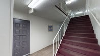 This interior common area serves as the central circulation space of a multi-unit building, offering a clean and functional layout designed to connect residents to their individual units. The ground-floor corridor stretches the length of the building, lined with light wood-look flooring that runs continuously underfoot, providing a neutral and durable surface throughout. Fluorescent lighting fixtures mounted to the textured ceiling ensure the hallway remains consistently well-lit from end to end.

Individual unit entry doors, finished in grey with raised panel detailing and brass numbering, are positioned at regular intervals along the corridor, beginning with unit 101. A staircase with dark burgundy carpeted treads and a white metal railing provides access to the upper floor, anchoring one end of the common area with a visual focal point. The open riser design and clean white balustrade contribute a straightforward, practical aesthetic.

A bank of grey metal mailboxes is conveniently located within the common area, offering secured mail access for multiple residents in one centralized location. Framed artwork displayed along the corridor walls adds a modest decorative touch, softening the utilitarian character of the space and creating a more welcoming atmosphere for residents and visitors alike.