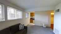 This single-floor space is thoughtfully laid out to maximize functionality within a compact footprint. The living area features neutral grey carpeting and white walls that create a clean, understated backdrop. A large window fitted with horizontal blinds allows generous natural light to filter in, offering a view of surrounding greenery and enhancing the sense of openness in the room.

The living area flows directly into the kitchen through an open doorway, creating a connected and accessible layout. The kitchen is equipped with warm wood-toned upper and lower cabinetry, providing ample storage. Appliances include a stove, microwave, and what appears to be a dishwasher or compact washer, all neatly arranged along the kitchen wall. The kitchen floor features light-toned tile, which contrasts with the carpeted living space and helps visually define the two areas.

The overall ambiance of the space is modest and functional, with a neutral color palette throughout. The open sightlines between rooms contribute to an airy feel despite the unit's compact scale. The space is well-suited for those seeking a straightforward, easy-to-maintain living environment.