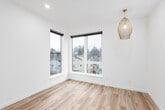This thoughtfully designed single-floor unit offers a clean, contemporary aesthetic throughout. The open-concept layout is anchored by light-toned hardwood flooring that extends seamlessly across the entire space, enhancing the sense of openness and continuity.

The main living area is flooded with natural light, courtesy of a striking set of large corner windows that frame views of the surrounding residential neighborhood. A woven rattan pendant light adds a warm, organic contrast to the otherwise crisp, white interior. Recessed lighting complements the natural light, ensuring the space remains bright at all hours.

The compact kitchenette is efficiently designed, featuring wood-toned lower cabinetry, a white countertop with an integrated sink, open floating shelves above, and space for essential appliances including a compact refrigerator. The layout maximizes functionality without sacrificing the open feel of the unit.

The walls throughout are finished in a clean, bright white, creating a versatile backdrop that enhances the sense of space. Additional windows offer views of the neighboring structures and greenery, further connecting the interior to its surroundings. The entry area includes a utility panel and additional storage provisions, ensuring practical needs are well accommodated. Overall, the unit presents a modern, minimalist character with warm natural accents.