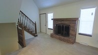 This three-story home showcases a blend of traditional architectural elements and practical design. The main living area is immediately notable for its vaulted ceiling, which lends a sense of openness and volume to the space. A full-height brick fireplace serves as the room's focal point, complete with a wooden mantel and a classic tri-panel screen, adding warmth and character to the interior.

Plush wall-to-wall carpeting in a neutral tone runs throughout the living area, contributing to a comfortable, understated aesthetic. A wooden staircase with turned spindle balusters and a solid newel post anchors one side of the room, providing access to the upper levels while adding a traditional, crafted detail to the space. Natural light enters through a window positioned near the fireplace, brightening the room without overwhelming the cozy atmosphere.

A doorway off the main living area provides additional access to adjoining spaces. The neutral wall color throughout complements the warm tones of the brick and wood elements, creating a cohesive and inviting environment. With three floors in total, the home offers ample vertical space and room for varied living arrangements across its levels.