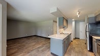This single-floor unit is designed with a practical, open-concept layout that connects the kitchen and living areas seamlessly. The kitchen is a standout feature, offering gray cabinetry, a white countertop peninsula with an integrated sink, and a full suite of appliances including a black electric range and stainless steel refrigerator. Track lighting illuminates the galley-style workspace, while wood-look plank flooring extends throughout the kitchen and living areas, adding warmth and durability.

The spacious living room benefits from natural light provided by a large double window and access to an outdoor balcony or patio, offering views of surrounding greenery. Neutral gray walls and a textured ceiling complete the area's clean, contemporary aesthetic.

Two bedrooms are finished with plush carpeting and gray walls. One bedroom includes a reach-in closet with built-in shelving, while the other features a mirrored sliding-door closet. Both rooms include windows that bring in natural light.

The bathroom is finished with wood-look tile flooring and includes a full-size soaking tub and shower combination, a vanity with gray cabinetry, and brushed nickel fixtures, providing a cohesive and functional design throughout.