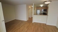 This single-floor space offers a clean, open-concept layout that integrates the living and kitchen areas into one cohesive environment. Warm-toned hardwood flooring runs throughout, lending a sense of continuity and a welcoming feel to the interior.

The kitchen is positioned along one side, anchored by a raised granite-topped breakfast bar that serves as a natural divider between the cooking and living zones. Cabinetry in a honey-maple finish provides ample storage, while stainless steel appliances — including a refrigerator, range, and over-the-range microwave — give the kitchen a modern, functional character. A stainless steel sink completes the workspace.

The open living area extends generously from the kitchen, offering flexible space for furnishing and everyday use. Neutral white walls and flush-mount ceiling lighting keep the space bright and clean throughout. A hallway visible toward the rear of the space leads to additional areas, including what appears to be a closet and a connecting room, suggesting thoughtful use of the overall floor plan.

The overall ambiance is practical yet inviting, with a neutral palette and consistent finishes that create a move-in-ready environment well-suited for comfortable daily living.