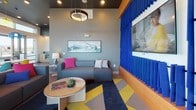 Located at 1493 Osage St in Denver's dynamic West Colfax neighborhood, this single-floor community lounge is a boldly designed, energetic space built for socializing and connection. The interior makes a strong design statement through a carefully curated palette of vivid colors and contemporary furnishings. A lounge area anchors one end of the space, centered around a plush gray sofa accented with bright pink throw pillows and a round wood coffee table, all set atop a graphic geometric area rug in navy, yellow, and pink. A striking blue vertical slat feature wall frames a large wall-mounted television, adding an eye-catching architectural focal point. Pendant lighting with circular fixtures and dark recessed ceiling details lend a sophisticated contrast overhead. The dining and co-working zone is bathed in natural light through expansive floor-to-ceiling windows that frame sweeping panoramic views of the Denver skyline. A long wooden counter with bar-height seating runs the length of the window wall, while a large communal table surrounded by colorful mixed chairs provides flexible dining or meeting space. A bold red pendant light overhead adds a playful pop of color. Large glass doors open directly to an outdoor terrace, seamlessly blending indoor and outdoor living. The overall ambiance is modern, lively, and thoughtfully designed to inspire community interaction.