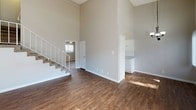 This two-story space makes an immediate impression with its dramatic double-height ceilings in the main living area, creating a sense of openness and volume that defines the overall ambiance. The floors throughout the main level are finished in warm, rich hardwood-style planks, adding both visual warmth and durability to the space. Neutral greige-toned walls provide a clean, versatile backdrop that complements a wide range of furnishing styles.

A carpeted staircase with a white railing leads to the upper level, positioned along one side of the living area. Adjacent to the staircase, a doorway opens into a secondary room on the main floor, which benefits from natural light filtering through a window. Partially visible from the living area, the kitchen features white cabinetry and appliances, offering a functional layout tucked neatly to one side. The open sightlines between the living and kitchen areas contribute to a connected, practical flow throughout the main level. Overall, the space balances volume and functionality in a clean, understated design.