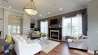 This single-floor communal lounge is thoughtfully designed to offer both comfort and sophistication. The main seating area centers around a striking fireplace with a bold black surround and cream-toned surround detail, above which a wall-mounted television is installed. The dark accent wall provides a dramatic backdrop that contrasts beautifully with the lighter gray tones found throughout the rest of the space.

Rich hardwood flooring runs throughout the primary lounge area, anchored by a neutral area rug beneath a round glass-top coffee table. Seating includes a white sofa, upholstered armchairs, and accent chairs with patterned fabrics, all accented with yellow throw pillows that add a lively pop of color. A decorative rope-wrapped pendant light fixture hangs overhead, adding a refined touch to the room's ambiance.

Large multi-pane windows along multiple walls flood the space with natural light and offer views to the outdoors. Crown molding and recessed lighting enhance the architectural detail of the vaulted ceiling. A bistro-style high-top table with bar chairs occupies a secondary nook off the main corridor. French doors visible in the background suggest an additional sunlit room or outdoor access, further expanding the open and airy feel of this well-appointed communal area.