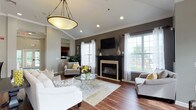 This single-floor communal lounge is thoughtfully designed to offer both comfort and sophistication. The main seating area centers around a striking fireplace with a bold black surround and cream-toned surround detail, above which a wall-mounted television is installed. The dark accent wall provides a dramatic backdrop that contrasts beautifully with the lighter gray tones found throughout the rest of the space.

Rich hardwood flooring runs throughout the primary lounge area, anchored by a neutral area rug beneath a round glass-top coffee table. Seating includes a white sofa, upholstered armchairs, and accent chairs with patterned fabrics, all accented with yellow throw pillows that add a lively pop of color. A decorative rope-wrapped pendant light fixture hangs overhead, adding a refined touch to the room's ambiance.

Large multi-pane windows along multiple walls flood the space with natural light and offer views to the outdoors. Crown molding and recessed lighting enhance the architectural detail of the vaulted ceiling. A bistro-style high-top table with bar chairs occupies a secondary nook off the main corridor. French doors visible in the background suggest an additional sunlit room or outdoor access, further expanding the open and airy feel of this well-appointed communal area.