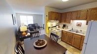 This single-floor unit is designed around an open-concept layout that seamlessly integrates the kitchen, dining area, and living room into one cohesive living space. The kitchen is equipped with warm-toned oak cabinetry offering generous upper and lower storage, a white electric range with an overhead ventilation hood, and a double-basin sink set into a continuous countertop. A patterned area rug anchors the kitchen floor, adding a decorative touch to the tile. Adjacent to the kitchen, the dining area centers on a dark-stained wood table with matching chairs, creating a defined yet open space for meals. A decorative bowl with pinecones serves as a centerpiece. The living room flows naturally from the dining space, featuring light-toned carpeting that contrasts the kitchen's tile flooring. The seating area includes a sofa and additional chairs arranged around a glass-top coffee table. A large window allows natural light to fill the living room, contributing to the bright and airy feel of the space. A floor lamp adds warm ambient lighting near the dining zone. The overall palette is neutral with yellow accent touches throughout, lending the space a welcoming and cohesive character.
