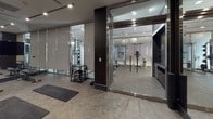 This contemporary fitness facility is thoughtfully designed to accommodate a wide range of training styles within a single, cohesive space. The layout is divided into distinct functional zones, each separated by full-height glass partitions and dark structural columns that lend a sophisticated, upscale aesthetic throughout.

The main exercise studio is a standout feature, lined floor-to-ceiling with mirrors that amplify the sense of space and allow for form monitoring during workouts. Recessed track lighting illuminates the room with a focused, atmospheric glow. The studio is equipped with stability balls, weight benches, balance discs, and yoga mats arranged neatly across the durable stone-finish flooring. A large flat-screen display mounted on a freestanding stand serves as a central focal point for guided training sessions or class instruction.

Adjacent to the studio, a glass-enclosed strength training room houses cable machine systems and suspension training equipment, offering a well-appointed space for resistance-based workouts. The open-plan functional training area near the entrance features plyo boxes, kettlebells, and additional floor mats, providing versatility for dynamic and conditioning exercises.

The overall design palette of charcoal, dark wood, and polished concrete creates a refined, high-performance environment that balances functionality with a distinctly premium atmosphere.