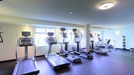 This fitness center is thoughtfully designed to support an effective workout experience. The space is lined with a series of large windows that allow natural light to fill the room, creating a bright and energizing atmosphere. A row of modern cardio machines — including treadmills and elliptical trainers, each equipped with individual display screens — is positioned along the windowed wall, allowing users to enjoy outdoor views while exercising. The flooring is a durable dark rubber material, well-suited for high-traffic fitness use. Ceiling-mounted light fixtures provide consistent overhead illumination throughout the space. Structural columns are integrated cleanly into the layout, and a mirrored wall along one side enhances the sense of openness and allows users to monitor their form. The overall design is clean, functional, and modern, with a neutral color palette of whites and grays that contributes to a focused and professional environment. This single-floor facility is well-organized and offers a comfortable setting for cardiovascular training.