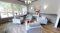 Located at 3800 14th Ave SE in Lacey, Washington, this single-floor rental office is thoughtfully designed to create a warm and professional first impression for prospective residents. Upon entry, a bold black double front door with transom windows and sidelights sets an elegant tone, flooding the foyer with natural light and offering views of the surrounding greenery. A standout rustic barn door with richly textured wood paneling slides along a black iron track, serving as both a functional divider and a striking design feature that bridges modern and industrial aesthetics.

The main waiting area is furnished with contemporary light gray upholstered armchairs arranged around natural wood stump accent tables, anchored by warm hardwood-style flooring throughout. A multi-arm chandelier with frosted cylindrical shades illuminates the room, complemented by wall sconces and vibrant geometric artwork that adds a pop of color to the neutral gray palette. Large windows frame views of mature trees outside, enhancing the bright and airy atmosphere.

A dedicated leasing counter with a dark countertop and modern white bar stools provides a functional workspace for staff-resident interactions, with a pass-through window and access to a back office area. Crown molding and a two-tone wall treatment with a charcoal chair rail add architectural polish throughout the space.