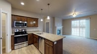 This single-floor home offers a thoughtfully designed open-concept layout that seamlessly connects the kitchen and living area. The kitchen is a standout feature, showcasing rich dark wood cabinetry, granite countertops, a central island with a built-in sink, stainless steel appliances — including a refrigerator, range, microwave, and dishwasher — and elegant pendant lighting overhead. Light wood-style flooring runs throughout the kitchen, transitioning to plush carpet in the living area. The living room is anchored by a classic stone-surround fireplace with a white mantel, a ceiling fan, and access to a sliding glass door that brings in natural light and outdoor views. The bedrooms are generously sized, each featuring soft carpet flooring, ceiling fans, and windows that allow ample natural light. One bedroom includes a vaulted ceiling, adding a sense of openness. The bathrooms are finished with warm wood vanities, white countertops, and durable wood-look vinyl flooring. A second bathroom features grab bars and additional accessibility accommodations. Neutral tones throughout the home create a clean, move-in-ready aesthetic that is both functional and inviting.