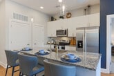 Located at 2–14 S Clayton St in Lawrenceville, GA, this thoughtfully designed single-floor residence offers a cohesive blend of modern finishes and functional living spaces. The open kitchen is a standout feature, showcasing granite countertops, white shaker cabinetry extending to the ceiling, stainless steel appliances including a range, microwave, and refrigerator, and a peninsula with bar seating — all set atop warm wood-look flooring. The living area is bright and welcoming, with large sliding glass doors leading to a private balcony that frames outdoor views, complemented by a bold accent wall and stylish décor. Multiple bedrooms provide comfortable retreats, each featuring plush carpeting, generous windows with natural light, and ample space. The primary bedroom connects to a well-appointed en-suite bathroom with granite countertops, white cabinetry, brushed nickel fixtures, a framed mirror, and a glass-enclosed walk-in shower. A second bathroom mirrors the same quality finishes. Throughout the residence, a consistent neutral palette punctuated by contemporary accents creates a polished and inviting atmosphere well-suited for modern living in the heart of Lawrenceville.