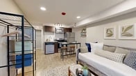 This thoughtfully designed studio maximizes every square foot with a smart, open-concept layout that seamlessly integrates the living and kitchen areas. Warm wood-look flooring runs throughout, complemented by neutral gray walls and clean white trim that create a bright, cohesive aesthetic. A ceiling fan with pendant lighting anchors the living area, which benefits from a window that allows natural light to filter in.

The kitchen is well-appointed, featuring dark espresso cabinetry, a gray subway tile backsplash, and a full suite of stainless steel appliances including a refrigerator, range, microwave, and dishwasher. A built-in bar-height dining table with industrial-style stools provides a practical and stylish dining solution adjacent to the kitchen. Track lighting overhead keeps the space well-lit and functional.

The bathroom includes a full soaking tub and shower combo with vertical gray tile detailing, dark wood vanity cabinetry, and clean white fixtures. The space is compact yet complete, offering all essential amenities. A wall-mounted HVAC unit in the living area ensures year-round comfort. Overall, this studio presents a modern, efficient living environment with carefully considered finishes throughout.