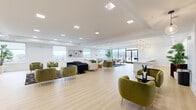 This elegantly designed single-floor amenity space seamlessly blends modern aesthetics with biophilic design elements. The entrance immediately impresses with a dramatic full-height living green wall, lush with layered foliage, which sets a vibrant, nature-inspired tone throughout. Light oak hardwood flooring extends across the entire space, complementing the crisp white walls and ceilings fitted with recessed square skylights that flood the interior with natural light.

The expansive lounge area is thoughtfully arranged with plush velvet seating in rich olive green, navy, and cream tones, creating intimate conversation zones across the open floor plan. A standout living wall in the main lounge also features an integrated fireplace and mounted display screen, serving as a bold focal point. Adjacent to this, a full bar and kitchen area showcases a striking white textured accent wall, dark wood cabinetry, open floating shelves, a generous white quartz countertop, and a row of navy and gold bar stools beneath sculptural geometric gold pendant chandeliers.

Panoramic windows throughout offer sweeping elevated views of the surrounding cityscape and hillside beyond. Floor-to-ceiling glass sliding doors connect the interior to a spacious outdoor terrace, extending the living area and enhancing the sense of openness. The overall ambiance is refined, airy, and welcoming.