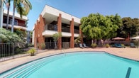This property showcases a striking multi-story architectural design that blends classic brick columns with smooth stucco facades, creating a warm and structured aesthetic. The exterior is defined by prominent red brick pillars that rise through each level, supporting covered balconies with wrought-iron railings that overlook the outdoor amenity area below.

The centerpiece of the outdoor space is a generously sized swimming pool with clear turquoise water, complemented by a circular spa situated directly adjacent. The pool deck is finished in light-toned concrete, providing ample space for lounging, and is furnished with reclining chairs positioned to take advantage of the open-air environment. A wrought-iron perimeter fence encloses the area, adding both security and a refined aesthetic boundary.

Mature trees, including a broad canopy tree and tall palms, provide natural shade and greenery throughout the courtyard, enhancing the resort-like atmosphere. Potted tropical plants are thoughtfully placed along the building's base, softening the transition between architecture and landscape. The overall ambiance is bright, open, and inviting, well-suited to warm-weather outdoor living.