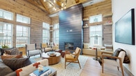 Located at 10952 Netherly Dr in Charlotte, NC, this single-story communal space masterfully blends rustic warmth with contemporary design. The architecture is defined by soaring vaulted ceilings supported by exposed timber trusses, horizontal wood-planked walls, and wide-plank hardwood flooring that together create a cohesive lodge-inspired aesthetic.

The main lounge area serves as the heart of the space, anchored by a dramatic floor-to-ceiling fireplace clad in deep blue-gray paneling that provides a striking modern contrast to the warm wood surroundings. Multiple seating arrangements, including mid-century style armchairs, plush sofas, and woven leather ottomans, are grouped around a circular wood coffee table, encouraging both relaxed conversation and casual gathering.

Natural light pours in through an abundance of windows, including transom windows positioned high along the walls, offering views to an outdoor pool and surrounding community. A cozy built-in window seat adds a charming nook for quieter moments.

A dedicated wet bar area features sleek cabinetry, a utility sink, and a beverage refrigerator, backed by an eye-catching mosaic tile wall in metallic blues and blacks. Intimate bistro-style seating tucked alongside the wood-planked walls adds additional flexible space throughout.