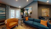 This thoughtfully curated single-floor space combines comfort and contemporary style across several distinct functional areas. The main lounge is anchored by vaulted ceilings that lend an open, airy atmosphere, complemented by deep slate-gray and teal-toned walls that create a cohesive, modern aesthetic throughout. Rich hardwood-style flooring runs continuously across the space, tying the areas together seamlessly.

The lounge area is well-appointed with a teal upholstered sofa, cognac leather accent chairs, and a glass-top coffee table, all arranged around a central seating arrangement. Large abstract paintings adorn the walls, adding artistic character, while floor-to-ceiling drape-framed doors allow natural light to filter in and provide views of the greenery outside.

A media and dining nook features a wall-mounted flat-screen television flanked by additional large-scale artwork, a mirrored console, and a glass bistro-height table with acrylic chairs — ideal for casual dining or group gatherings.

The full kitchen is equipped with stainless steel appliances, white shaker-style cabinetry, dark countertops, and a generous marble-topped island with bar seating, offering ample space for food preparation and entertaining.

A secondary kitchenette area includes white cabinetry, a utility sink, and a coffee station, providing convenient refreshment access. Throughout the space, carefully placed greenery and layered lighting enhance the welcoming and functional ambiance.