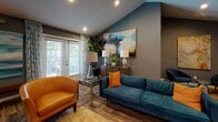 This thoughtfully curated single-floor space combines comfort and contemporary style across several distinct functional areas. The main lounge is anchored by vaulted ceilings that lend an open, airy atmosphere, complemented by deep slate-gray and teal-toned walls that create a cohesive, modern aesthetic throughout. Rich hardwood-style flooring runs continuously across the space, tying the areas together seamlessly.

The lounge area is well-appointed with a teal upholstered sofa, cognac leather accent chairs, and a glass-top coffee table, all arranged around a central seating arrangement. Large abstract paintings adorn the walls, adding artistic character, while floor-to-ceiling drape-framed doors allow natural light to filter in and provide views of the greenery outside.

A media and dining nook features a wall-mounted flat-screen television flanked by additional large-scale artwork, a mirrored console, and a glass bistro-height table with acrylic chairs — ideal for casual dining or group gatherings.

The full kitchen is equipped with stainless steel appliances, white shaker-style cabinetry, dark countertops, and a generous marble-topped island with bar seating, offering ample space for food preparation and entertaining.

A secondary kitchenette area includes white cabinetry, a utility sink, and a coffee station, providing convenient refreshment access. Throughout the space, carefully placed greenery and layered lighting enhance the welcoming and functional ambiance.