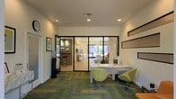 This single-story office space presents a clean, modern aesthetic with a well-organized layout designed for professional use. The main reception area features a sleek white desk accompanied by stylish lime-green accent chairs, creating an inviting and contemporary first impression. Underfoot, patterned carpet tiles in shades of green and gray add visual interest to the space while maintaining a professional atmosphere.

Recessed lighting throughout the ceiling provides consistent, even illumination across the workspace. Glass partitions with black metal framing divide the interior into distinct areas, allowing natural light to flow freely while maintaining defined spaces. Through the glass walls, a secondary room is visible, offering additional workspace and glimpses of the outdoors, suggesting strong natural light availability.

An adjacent room visible through a doorway on the left features framed artwork on the walls and a bar-height work surface with seating, functioning as a display or consultation area. The overall color palette of white walls, green furnishings, and natural light gives the space a fresh, professional ambiance suitable for client-facing business operations.