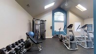 This fitness center is designed to support a comprehensive workout experience within a thoughtfully appointed single-level space. The room is defined by its dramatic vaulted ceiling, which adds a sense of openness and airiness throughout. A bold blue accent wall frames a large arched window at the far end, allowing natural light to fill the space and creating a visually striking focal point. A skylight overhead further enhances the bright, energizing atmosphere.

The rubber-floored workout area is outfitted with a range of equipment to accommodate both strength and cardio training. A multi-station cable machine anchors the strength training section, complemented by an adjustable workout bench and a full rack of dumbbells in varying weights, catering to users of all fitness levels. For cardiovascular exercise, the space includes an elliptical trainer, a stationary upright bike, and a stair climber, providing diverse options for aerobic workouts. A stability ball rounds out the functional training offerings.

The neutral beige walls, paired with the vibrant blue accent, create a motivating yet clean aesthetic. Ceiling fans provide additional air circulation, ensuring comfort during intense workouts. Overall, this fitness center combines practical equipment with an inviting, well-lit environment.
