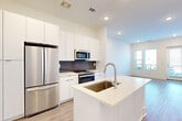 This well-appointed 1-bedroom, 1-bathroom residence offers a thoughtfully designed single-level layout that maximizes both function and comfort. The open-concept living and kitchen area is anchored by a sleek kitchen featuring floor-to-ceiling white cabinetry, a dark subway tile backsplash, stainless steel appliances including a French door refrigerator, microwave, and range, and a central island with a light quartz countertop and undermount sink. Light wood-look flooring runs throughout the main living areas, enhancing the bright, airy atmosphere created by recessed lighting and large windows. The living room opens directly to a private balcony through a glass door, allowing natural light to pour in and providing outdoor views. The bedroom is a comfortable, carpeted retreat with dual windows that frame exterior views and include a ceiling fan fixture. It connects conveniently to a dedicated closet and the full bathroom, which features white subway tile surround, a soaking tub, a vanity, and in-unit stacked washer and dryer. The overall design palette of crisp whites and neutral tones gives the space a clean, modern aesthetic throughout.