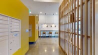 Located at 3836 Bank St in Baltimore, MD, this thoughtfully designed single-floor amenity space combines contemporary aesthetics with functional communal areas. The entrance corridor immediately establishes the design language with polished concrete floors, bold yellow accent walls, and a slatted wood partition that creates a sense of delineation without sacrificing openness. A built-in counter with bar-height seating and pendant lighting lines one wall, accompanied by framed abstract artwork and clear directional signage pointing residents toward the elevator, parking, and lounge.

The lounge area is a standout feature, boasting soaring ceilings and expansive floor-to-ceiling windows with dark metal frames that flood the space with natural light and offer views of an outdoor courtyard. Comfortable sectional seating, accent chairs, and a geometric gold chandelier contribute to an inviting, upscale atmosphere. A secondary lounge includes a slatted wood feature wall with integrated shelving and a television, anchored by a solid wood coffee table and plush seating.

The communal kitchen is well-appointed with teal cabinetry, white subway tile backsplash, stainless steel appliances, and a large terrazzo-topped island with bar seating—ideal for social gatherings. Soaring ceilings throughout reinforce the spacious, airy character of the entire amenity level.