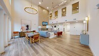 This expansive single-floor lounge and gathering space is defined by its dramatic double-height ceilings, which lend an open, airy quality to the entire interior. Wood-look tile flooring in warm, neutral tones runs throughout, unifying the various functional zones within the space. Two oversized circular brass chandeliers hang at staggered heights, serving as striking focal points and complementing the overall modern-transitional design aesthetic.

The lounge area centers on a large tile-clad accent wall featuring a sleek, linear gas fireplace and a wall-mounted flat-screen television. Seating consists of a gray sectional sofa, a pair of cognac leather accent chairs, and a low white coffee table, all arranged atop a neutral area rug. Adjacent to the lounge, a built-in shelving unit provides display and storage space. A secondary seating vignette nearby offers additional flexibility for gatherings.

The dining zone is positioned near a full wall of French doors and floor-to-ceiling windows that flood the space with natural light and provide direct views of an outdoor courtyard. A concrete-top dining table surrounded by woven rattan chairs completes this area. A welcoming entry foyer with a console table, curated artwork, and wall sconces provides an elegant transition into the space. Additional corridors lead to further amenities beyond.