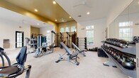 This single-floor fitness center is designed to deliver a full-scale workout experience in a well-appointed, open environment. The space features soaring vaulted ceilings with recessed lighting and dual ceiling fans, creating an airy and comfortable atmosphere. Large multi-pane windows line the walls, flooding the interior with natural light and offering views of the surrounding greenery.

The strength training area is extensively equipped, including a Matrix functional trainer with a full array of cable attachments, a multi-station cable machine, adjustable weight benches, a power rack, and a wall-mounted ladder rack. A dedicated dumbbell section features a complete rack of weights alongside a full set of kettlebells displayed on tiered storage racks, complemented by a large wall mirror to assist with form and technique.

The cardio zone is equally well-stocked, housing Matrix-brand treadmills, stationary cycling bikes, and an elliptical trainer, all positioned near the windows to maximize natural light during workouts. The neutral-toned carpet flooring, warm gold accent walls, and crisp white trim create a clean, motivating aesthetic throughout. A water fountain station and direct exterior access add practical convenience to this comprehensive fitness facility.