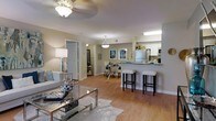 This single-floor residence showcases an open-concept design that effortlessly connects the living, dining, and kitchen areas into one cohesive, flowing space. The living area features warm hardwood-style flooring, a ceiling fan with a light fixture, and ample room for comfortable furnishings, anchored by a glass-top coffee table and a plush area rug. A floor lamp adds a warm, ambient glow to the seating area.

The kitchen is positioned along one side and features white cabinetry, stone-look countertops, and stainless steel appliances, including a full-size refrigerator. A breakfast bar with white bar stools serves as both a functional prep space and a casual dining option, illuminated by a pendant light above.

Adjacent to the kitchen, a dedicated dining area accommodates a glass dining table with seating for several guests, complemented by decorative wall art that adds a coastal aesthetic. The layout allows natural sightlines throughout, creating an airy, open ambiance. A hallway visible from the main living area leads to additional rooms, suggesting a practical and well-organized floor plan throughout.