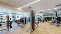 This fitness center is a thoughtfully designed, single-floor space that combines functionality with contemporary style. The flooring throughout is a standout feature — light-toned wood in a distinctive herringbone pattern that lends warmth and visual interest to the entire room. Recessed linear lighting runs along the drop ceiling, providing consistent, bright illumination across all workout zones.

The space is divided into distinct activity areas. A cardio section near the exterior windows houses Life Fitness upright bikes, ellipticals, and treadmills, benefiting from natural light and outdoor views. The strength training area is generously stocked with Life Fitness selectorized machines and a multi-functional cable station, supported by mirrored walls that enhance the sense of depth and allow users to monitor form.

A dedicated free weights zone offers a full dumbbell rack, benches, and a power rack. One wall features a bold geometric triangle-patterned accent in white and navy, paired with warm wood cabinetry, yoga mat storage, and wall-mounted flat-screen televisions, adding an entertainment element. Teal accent walls and large windows contribute to an open, energizing ambiance throughout.