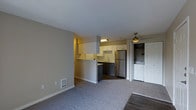 This single-floor space features an open-concept layout that seamlessly connects the living area to the kitchen. The living space is finished with plush grey carpeting, neutral-toned walls, and a ceiling fan overhead, contributing to a comfortable and functional atmosphere. A wall-mounted heating unit is conveniently positioned along the baseboard.

The kitchen is defined by a peninsula-style half-wall that creates a natural division between cooking and living areas while maintaining an open feel. White upper cabinetry lines the kitchen walls, complemented by dark lower cabinets and a stainless steel refrigerator. A stainless steel sink is positioned beneath under-cabinet lighting, and the kitchen flooring transitions to hardwood-style planks, offering a practical contrast to the carpeted living space.

Adjacent to the kitchen, a closeted laundry area houses a washer and dryer unit, adding everyday convenience. A bi-fold door provides easy access while keeping the area neatly tucked away. The overall design prioritizes efficient use of space, with a clean, neutral palette throughout that creates a bright and livable environment.