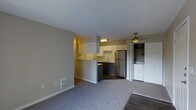 This single-floor space features an open-concept layout that seamlessly connects the living area to the kitchen. The living space is finished with plush grey carpeting, neutral-toned walls, and a ceiling fan overhead, contributing to a comfortable and functional atmosphere. A wall-mounted heating unit is conveniently positioned along the baseboard.

The kitchen is defined by a peninsula-style half-wall that creates a natural division between cooking and living areas while maintaining an open feel. White upper cabinetry lines the kitchen walls, complemented by dark lower cabinets and a stainless steel refrigerator. A stainless steel sink is positioned beneath under-cabinet lighting, and the kitchen flooring transitions to hardwood-style planks, offering a practical contrast to the carpeted living space.

Adjacent to the kitchen, a closeted laundry area houses a washer and dryer unit, adding everyday convenience. A bi-fold door provides easy access while keeping the area neatly tucked away. The overall design prioritizes efficient use of space, with a clean, neutral palette throughout that creates a bright and livable environment.