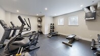 Located at 62 Armstrong Court in Halifax, Nova Scotia, this fitness center offers a well-appointed exercise room designed to support a comprehensive workout routine. The space features a clean, modern aesthetic with neutral-toned walls, recessed ceiling lighting, and durable rubber tile flooring throughout — ideal for both heavy equipment and high-intensity training.

The cardio section is equipped with multiple treadmills, elliptical trainers, and a stationary bike, providing ample options for cardiovascular exercise. A dedicated free weights area includes a multi-tiered dumbbell rack stocked with a full range of weights, accompanied by adjustable workout benches to support strength training. Stability balls are neatly stored on a vertical rack, and a battle rope is available for functional fitness training.

Climate comfort is maintained via a wall-mounted mini-split air conditioning unit, ensuring a comfortable environment year-round. A window along one wall allows for natural light, adding brightness to the space. The room is accessed through a solid dark-framed door, and the overall layout is thoughtfully organized to allow for safe movement between equipment zones. This fitness center provides residents with a convenient and fully functional on-site facility for their health and wellness needs.