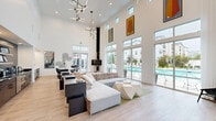 Located at 1100 Vintage Ln in Morgan Hill, California, this single-story community clubhouse is a standout example of contemporary design built for both comfort and functionality. The space is anchored by a dramatic great room boasting soaring double-height ceilings and walls of floor-to-ceiling glass that flood the interior with abundant natural light while framing direct views of the resort-style outdoor pool. Striking geometric pendant light fixtures add a sculptural element overhead, complementing the clean, white palette and light hardwood-style flooring that runs throughout. The main lounge area is generously proportioned, featuring a large sectional sofa, angular coffee tables, and curated modern artwork that lend the space a polished, upscale ambiance. A dedicated dining nook with a round white table and sleek black chairs provides a comfortable gathering spot. The kitchenette area is finished with bold dark cabinetry, dramatic veined black stone countertops, floating wood shelves, and stainless steel appliances, as well as a green tile backsplash accent wall that adds warmth and texture. The entryway greets visitors with soaring ceilings, glass panel doors, and a welcoming First Pointe Management Group branded mat, setting a professional tone. Together, these elements create an inviting, resort-caliber environment ideal for community gatherings.
