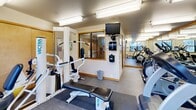 Located at 1040 NW 10th Ave in Portland's vibrant Pearl District, this fitness center is a well-equipped exercise room designed to support a complete workout experience within a compact, efficiently organized footprint. The space is outfitted with a diverse selection of cardio machines, including treadmills, ellipticals, and stationary bikes from brands such as True and Vectra, along with a multi-use strength training station and a workout bench. Large floor-to-ceiling mirrors line the walls, visually expanding the room and allowing users to monitor their form during exercise. Overhead fluorescent lighting ensures the space is bright and consistently illuminated throughout. Multiple windows along the exterior wall invite natural light and offer views of the surrounding Portland streetscape, adding an open, connected feel to the interior. The flooring is covered in durable, low-pile carpet with rubber mat sections beneath the equipment for stability and protection. A wall-mounted television provides entertainment during workouts, while a sanitizing station and posted informational signage support cleanliness and user guidance. Warm wood-toned trim accents the mirrors and doorframes, lending a polished finish to the otherwise functional design. The room is accessed through natural wood-finished doors, completing a clean and practical aesthetic throughout.